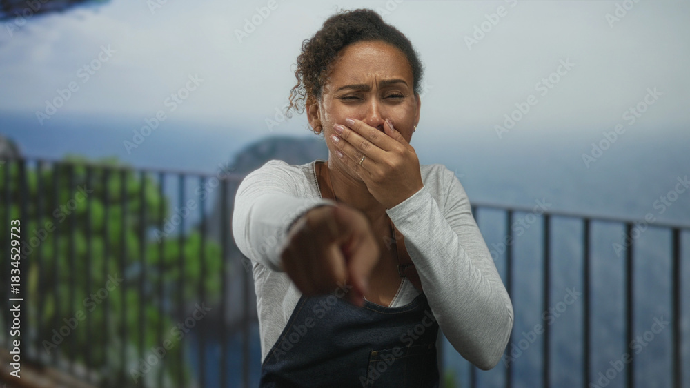 Fototapeta premium Woman covering mouth with hand and pointing finger toward camera at seaside railing; shock reaction.