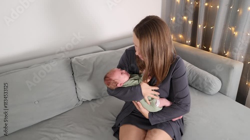 A woman is playing with a baby on a gray sofa in a room. The mother is rocking the crying baby in her arms. The girl is soothing the baby.