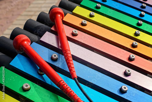 Foto A vibrant close-up of a colorful outdoor xylophone with two bright red mallets resting on the rainbow-colored keys in a public park, emphasizing musical play and childhood fun