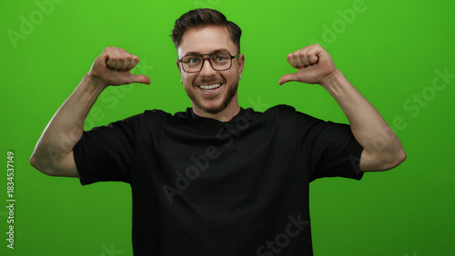 Caucasian man with glasses and beard pointing at himself against a bright green wall background wearing a black shirt and smiling confidently.