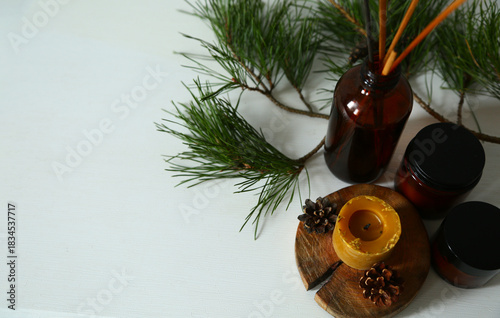 Wax candle on a wooden candlestick with green Christmas fir tree branch, brown aroma candles, aroma sticks in dark glass on a table. Ornament holiday background with white copy space. Natural light