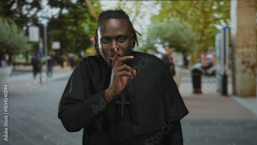 Young man priest wearing clerical collar and cross necklace, finger to lips for silence while standing on a street lined with trees; contemplation.