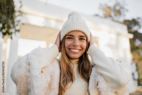 attractive smiling young blond woman walking in winter park having fun in warm white knitted sweater, cape and hat, cold season
