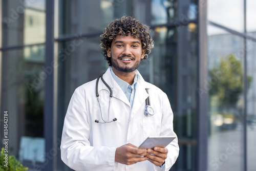 Slika na platnu Portrait of a young smiling Indian male doctor in a white coat standing outside,