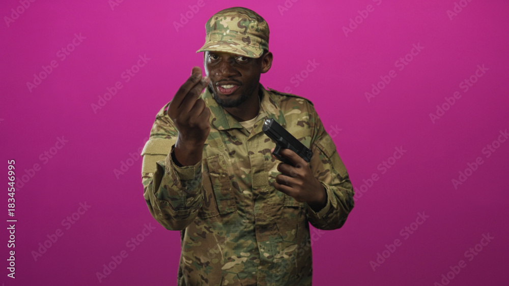 Fototapeta premium Man soldier in camouflage uniform holding a gun to his head while making a pinching fingers gesture against a magenta studio backdrop, smiling; tension threat.