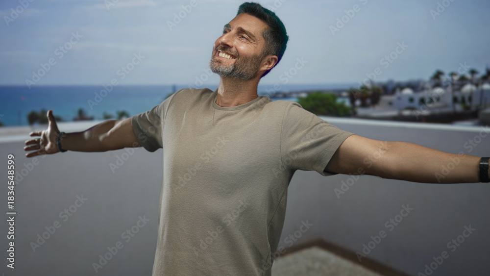 Fototapeta premium Man with arms outstretched showing hands on building rooftop terrace overlooking sea, smiling and looking up; joy escape calm.