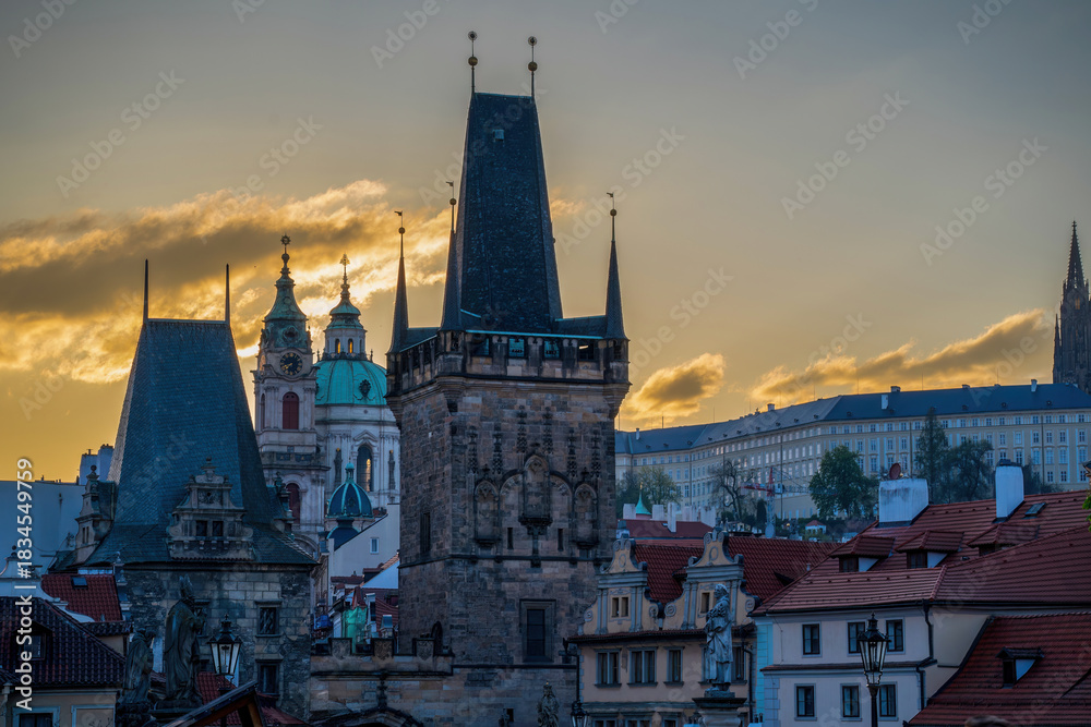 Fototapeta premium Sunset view of historic Prague with the Charles Bridge tower, old rooftops, and church domes glowing in warm evening light.