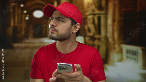 Фотография Young hispanic delivery man taps smartphone screen with focused gaze in church interior; contemplation faith devotion