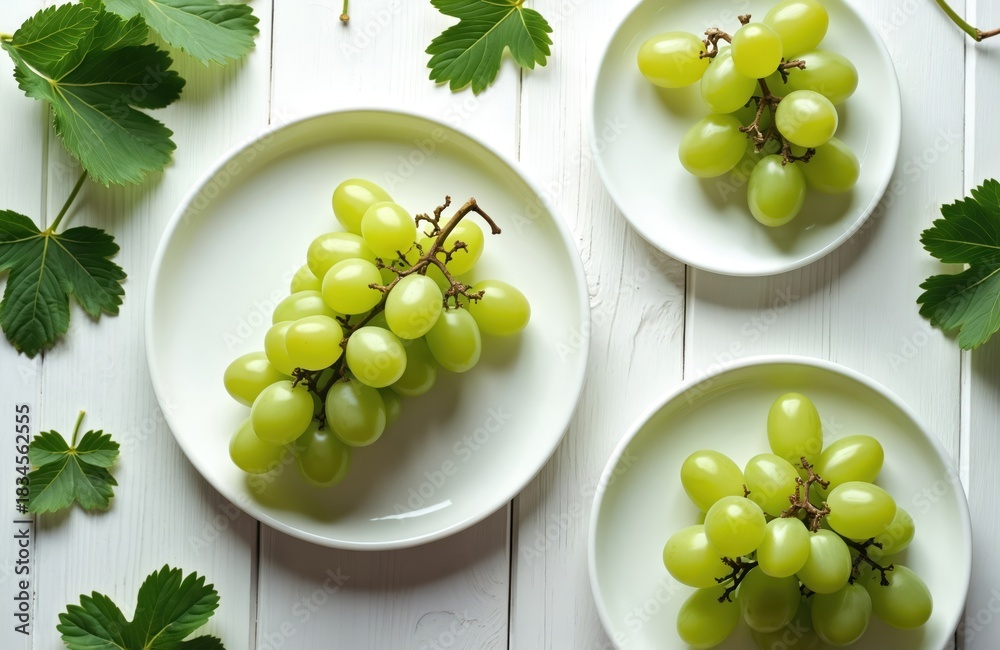 Obraz premium Clusters of fresh green grapes rest on white plates arranged on rustic wooden surface. Leafy green vines add natural touch to simple food composition. Shot perfect for healthy eating, farm to table