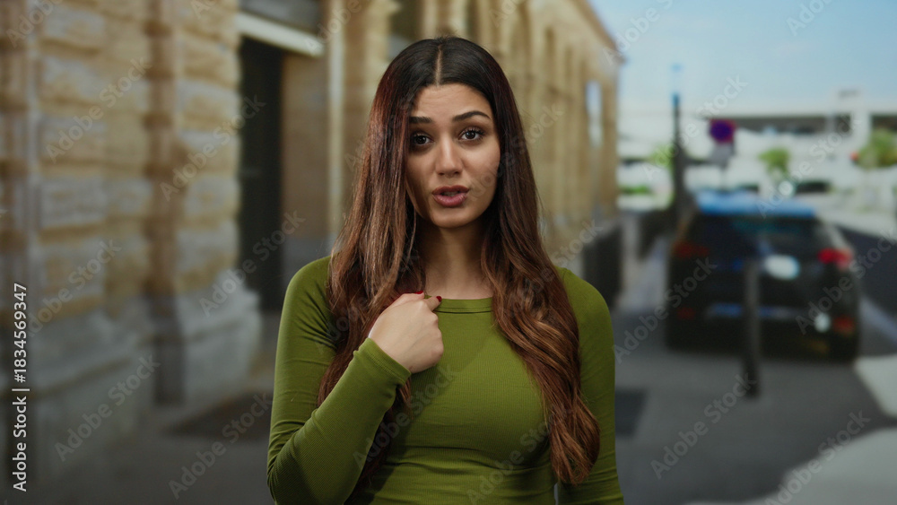 Fototapeta premium Woman outdoors gestures no with finger in urban street scene, exuding calm confidence, wearing green, hispanic descent, showcasing casual fashion against blurred city background.