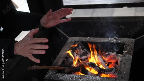 A man warming their hands over a small open fire at a Christmas market. The glowing flames create a cozy contrast to the cold winter atmosphere surrounding the festive outdoor event