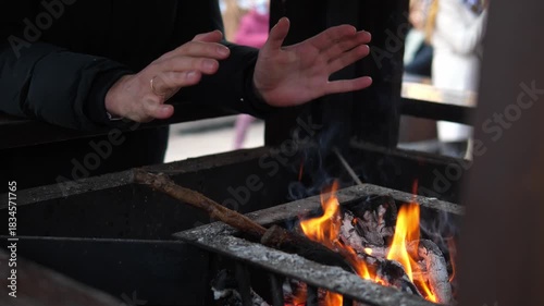 A man warming their hands over a small open fire at a Christmas market. The glowing flames create a cozy contrast to the cold winter atmosphere surrounding the festive outdoor event