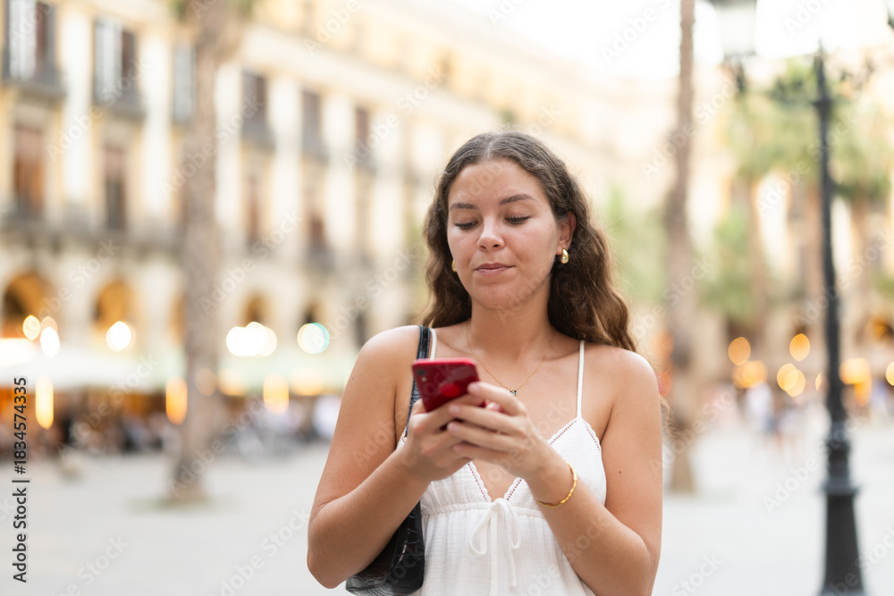 Obraz premium Young smiling woman with smartphone walks along Royal square, walks around city, spend time in public urban space. Electric street lighting, twilight in southern city, evening life in big city ..