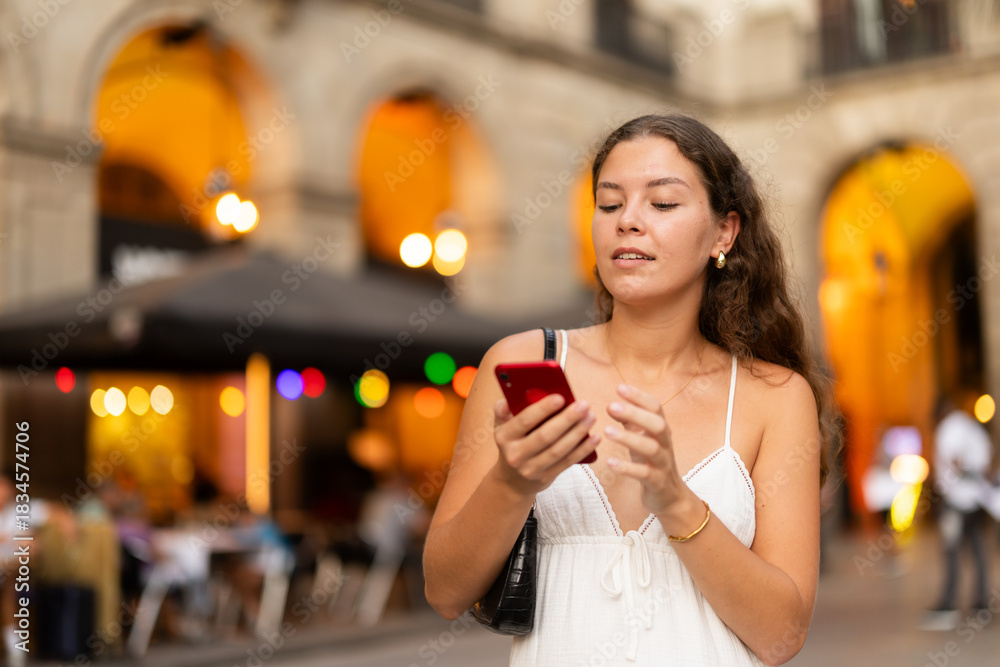 Obraz premium Young smiling woman with smartphone walks along Royal square, walks around city, spend time in public urban space. Electric street lighting, twilight in southern city, evening life in big city ..