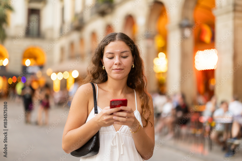 Obraz premium Long-haired cheerful female brunette walks along city street with mobile phone, spends time in motion, walks and enjoys sunny warm day, walk from university to home. Carefree, evening cafe, promenade