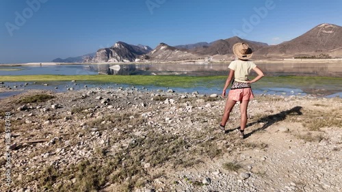 Woman in a cowboy hat relaxing on rugged rocks, enjoying the blue sky and turquoise water of the sea on Mughsayl Beach close to Salalah in Dhofar Governorate of Oman in dry season.