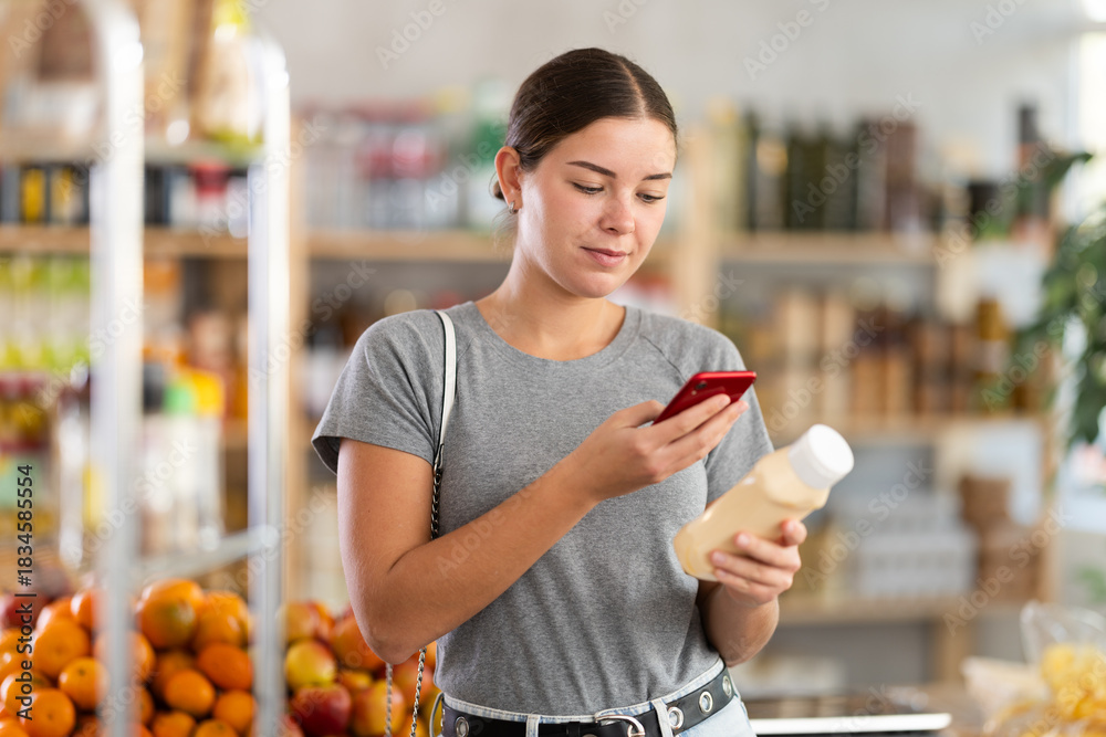 Fototapeta premium Young woman scans the barcode of a mayonnaise package on mobile phone. Visitor to a European store uses a smartphone to scan a QR code