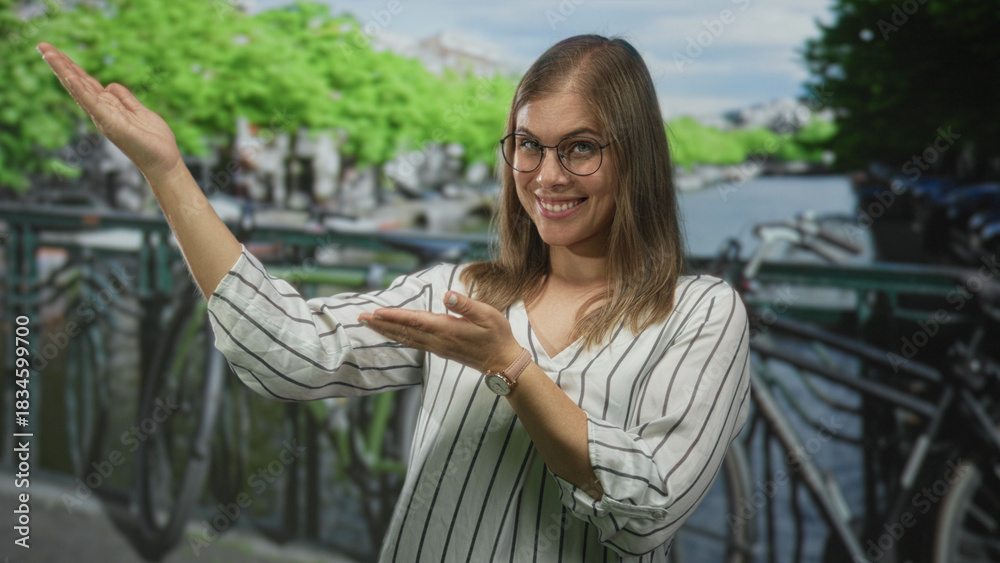 Fototapeta premium Woman extends hands to side on a street beside an amsterdam canal, smiling and gesturing palm up; joy welcome.