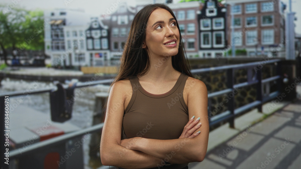 Fototapeta premium Woman with arms crossed showing bare shoulders standing on street in amsterdam by canal bridge and railing; confidence independence selfreliance.
