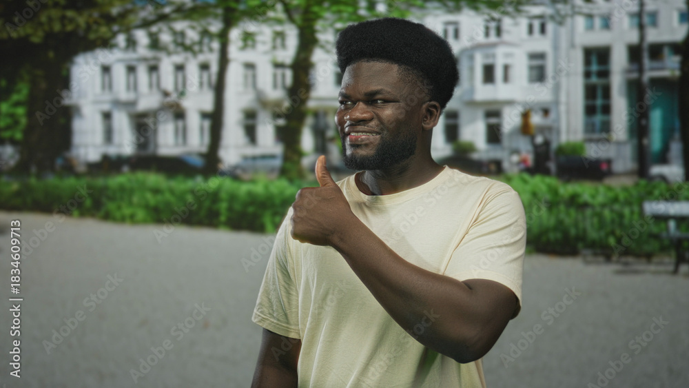 Fototapeta premium Man showing thumbs up on street in front of white apartment building and green park hedges, smiling with bare hand and thumb visible; confidence positivity.