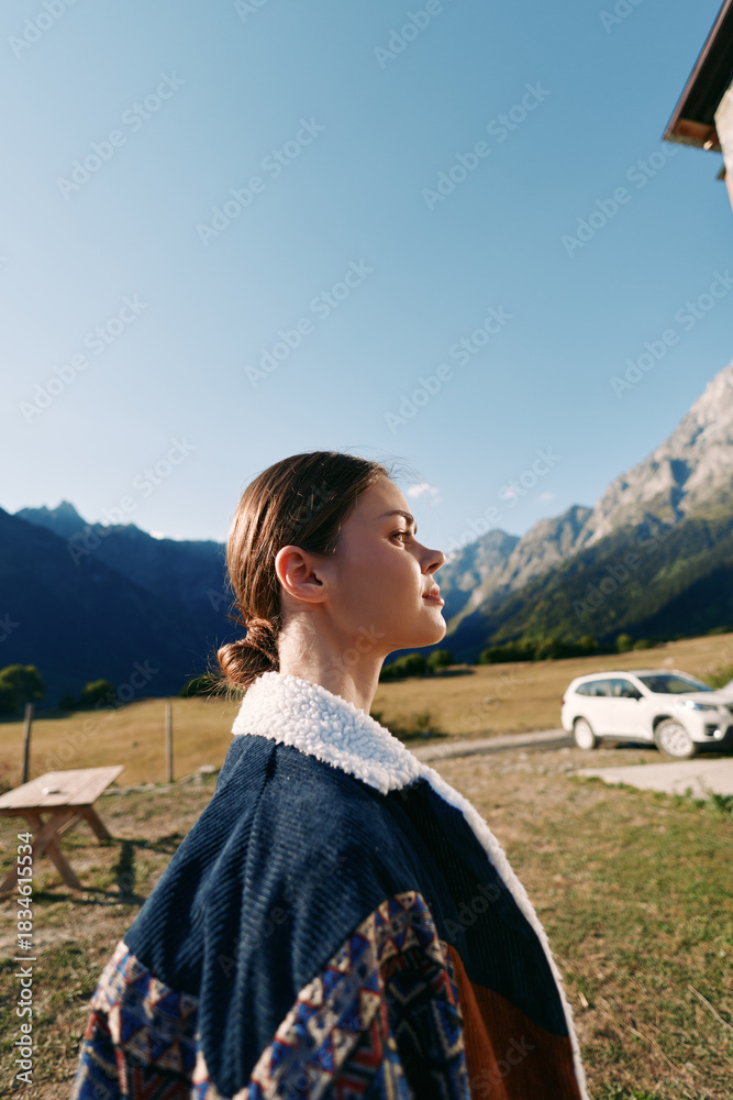 Naklejka premium Woman profile in mountains field by a roadside car, wearing a warm jacket and feeling sunlight on her face, outdoor portrait of a young traveler standing near picnic table under clear sky