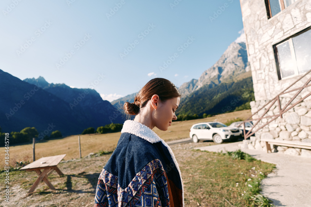 Fototapeta premium Woman standing near a stone chalet by a parked car in a grassy meadow, serene mountains in background. Outdoor portrait, travel lifestyle and rural countryside scene.
