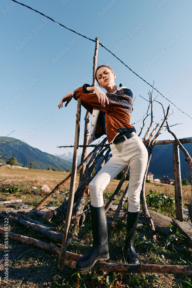 Naklejka premium Woman model in countryside wearing sweater and rubber boots, striking a confident pose while leaning on rustic fence with mountains and open field in natural sunlight.