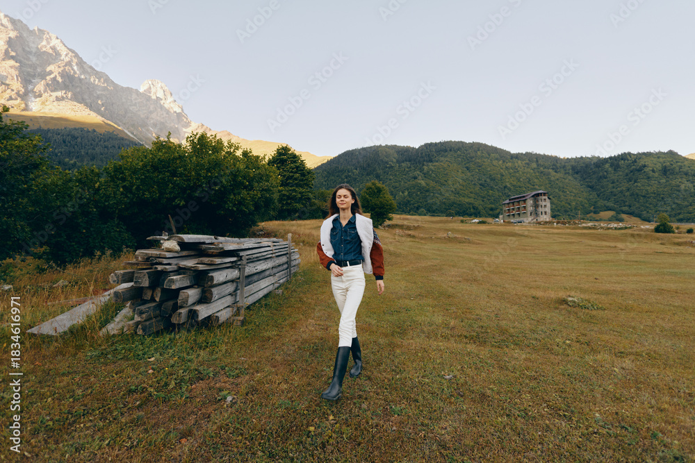 Fototapeta premium Woman walking in a grassy field near mountains and a wood pile, countryside scene with jacket and boots, autumn light and scenic nature for travel and outdoor lifestyle.
