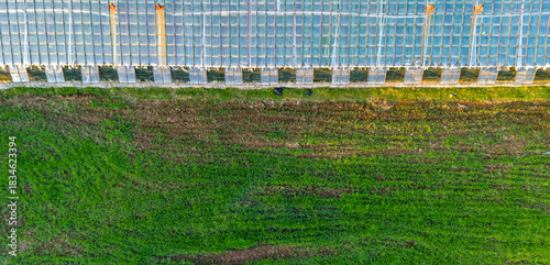 Top Down View Of The Edge Of A Glass Or Plastic Greenhouse Structure Bordering A Lush Green Grass Field In A Rural Farming Environment