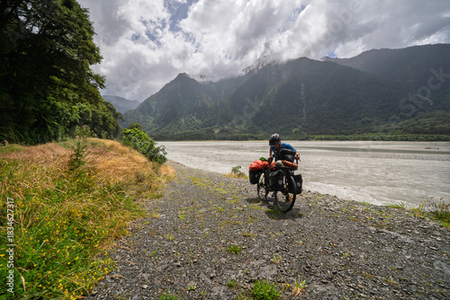 Male cyclist standing with loaded touring bike on gravel riverbed, New Zealand