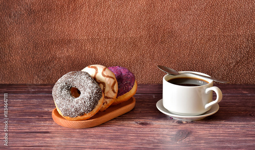 A cup of black coffee on a saucer with a spoon and a plate with three fresh donuts in multi-colored glaze on a wooden table.