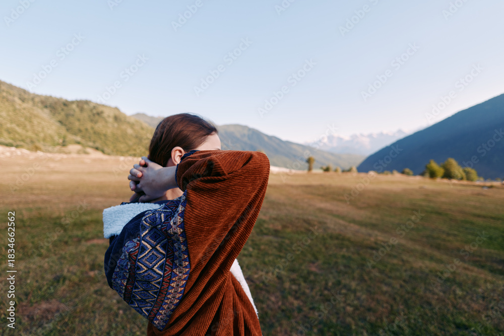 Fototapeta premium Woman in patterned sweater stretches and relaxes in nature on a grassy meadow with mountain landscape in the background, outdoors travel portrait capturing calm and freedom.