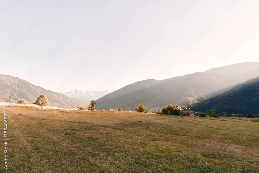 Fototapeta premium Meadow valley mountains grass landscape hills countryside rural scenery with open field, scattered trees and distant village under soft morning light and clear sky.