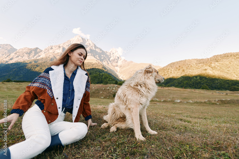 Fototapeta premium Woman and dog sitting in a mountain meadow outdoors, casual autumn jacket, enjoying nature and pet companionship, relaxed portrait with scenic peaks and open grassy field.