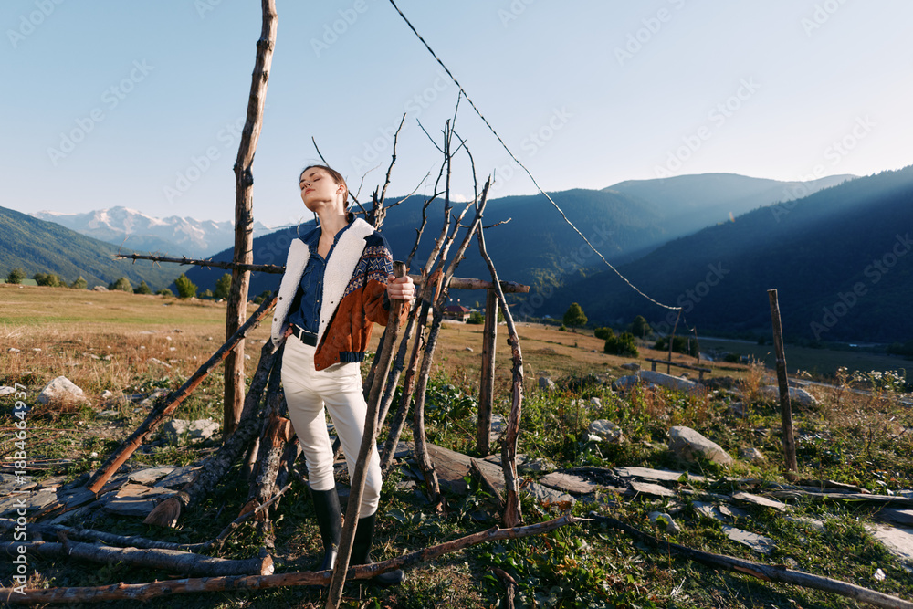 Fototapeta premium Woman in countryside near rustic fence and meadow, portrait enjoying sunlight with mountains in background and wide open field. Outdoor rural lifestyle, fashion and peaceful nature scene.