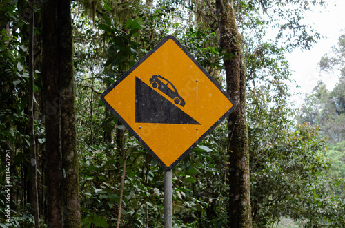 Bright yellow warning sign indicating steep downhill slope caution for drivers in forest