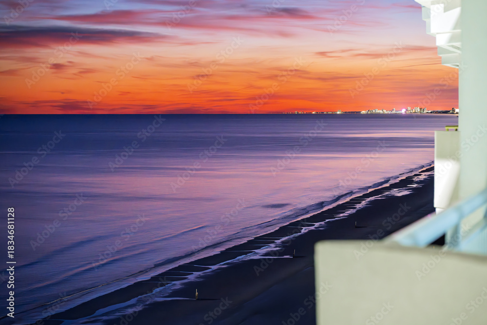 Fototapeta premium Balcony view of the beach and dunes under a colorful sunset with the illuminated downtown skyline in the distance, along the Grand Strand of the Atlantic Ocean at North Myrtle Beach, South Carolina.