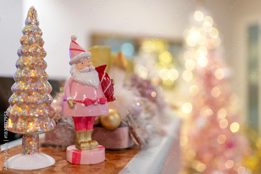 Fototapeta premium A pastel Christmas decoration scene featuring a pink Santa figurine and a glittery tree, captured with soft golden bokeh lights. Warm, festive, and perfect for holiday-themed designs.