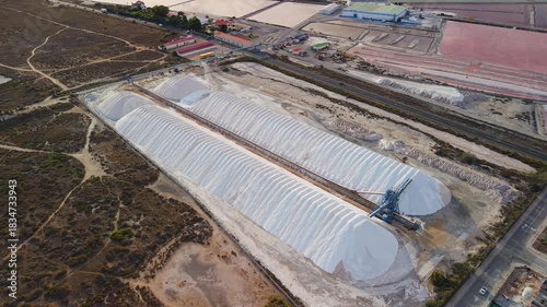 Aerial drone view of large salt piles at a production facility near Santa Pola, Spain, with colorful evaporation ponds and surrounding landscape.