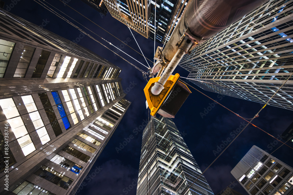 Fototapeta premium Night view looking up at illuminated skyscrapers with overhead transit wires and a traffic signal.