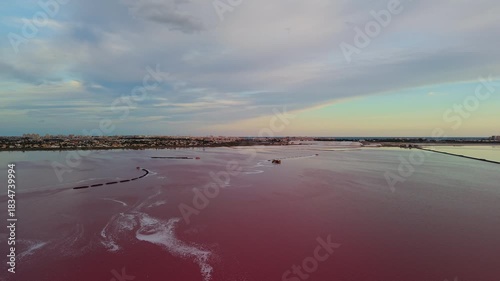 Aerial view of the pink salt lake Laguna Salada de Torrevieja in Spain at sunset, with salt extraction barges and the coastal city visible in the distance.