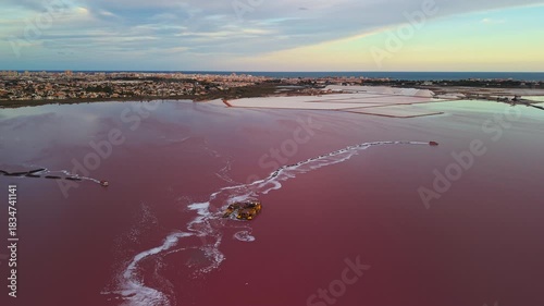 Aerial view of salt extraction barges operating on the pink waters of Laguna Salada de Torrevieja, Spain, with the city and salt mounds visible under colorful evening light.