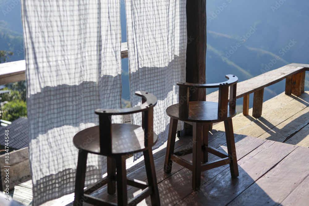 Obraz premium Two empty wooden chairs on rustic porch create relaxing and peaceful outdoor scene. Sunlight shines on simple furniture and white curtain, perfect for calm morning at cafe