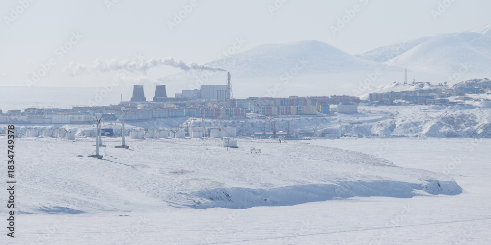 Fototapeta premium Winter Arctic landscape. View of the northern city. Anadyr, Chukotka, Russia. In the foreground are wind turbines and large fuel tanks at Cape Observation. Snowy tundra on the coast of a frozen river.