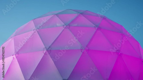 Abstract Geometric Dome Structure Illuminated with Vibrant Pink and Purple Hues Against a Clear Blue Sky.