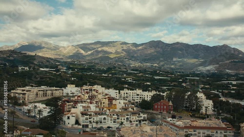 High-angle view from Salobreña castle showing white Andalusian town, rural valley and majestic Sierra Nevada mountains under clouds.