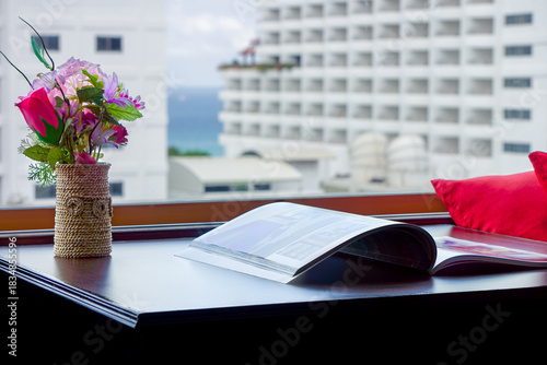 Window View with Magazine and Flowers in a Modern Hotel Room Setting. Dark wooden desk with a vase of artificial flowers and an open magazine, overlooking a high-rise building and the distant sea.