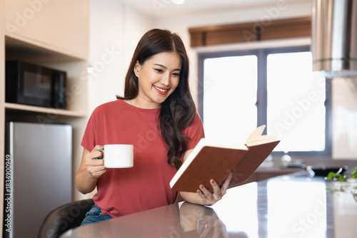 Pretty young woman with cup of coffee or tea reading book in modern kitchen counter at home apartment.  Morning lifestyle concept.