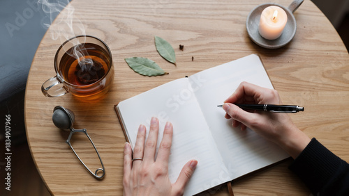 Woman hand writing in open notebook on wooden table. Cozy home work or study with tea and candle. Personal journal or creative writing concept.