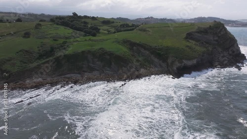 Side and upward drone shot on the Cantabrian coast, showing consecutive waves, bright green headlands, we see the town of Comillas in the distance, the sunlight reflecting softly on the sea.